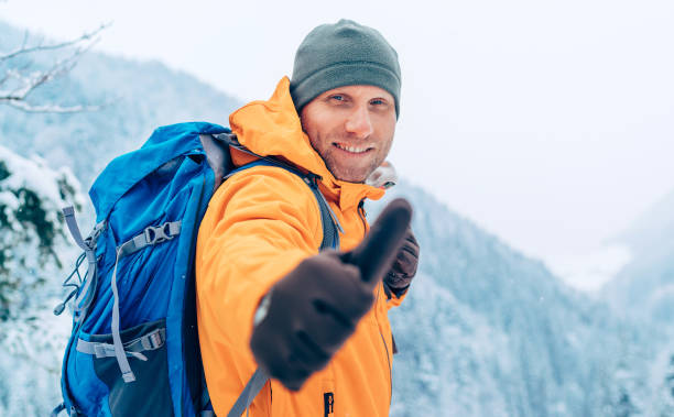 Smiling Man dressed bright orange softshell jacket with backpack looking at camera and showing a thumbs-up gesture while he trekking winter mountains route. Active people in the nature concept image. stock photo