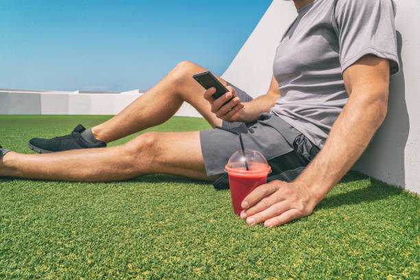Healthy active man using phone drinking beet juice smoothie drink takeaway cup on summer outdoor gym park background. Closeup of smartphone and plastic glass.