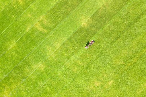 Aerial view of small figure of man worker trimming green grass with mowing mashine on football stadium field in summer.