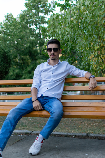 Stock photography portrait of a young man in the park, sitting on the park bench, with sunglasses, enjoying nice summer and sunny day, close up, smiling, looking at the view and camera.