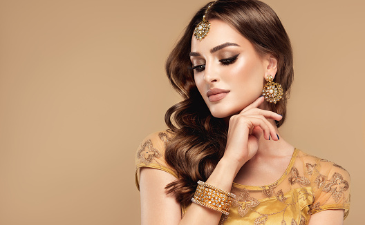 Young, brown haired woman with closed eyes is showing perfect makeup in Indian style on the flawless face. Gilded, hand made earrings and head adornment on her forehead. Woman beauty.