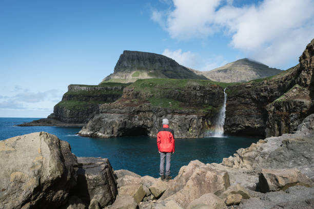 A tourist looks at a Mulafossur waterfall in Gasadalur on Faroe Islands A tourist looks at a Mulafossur waterfall in Gasadalur village. Vagar Island, Faroe Islands, Denmark vágar photos stock pictures, royalty-free photos & images