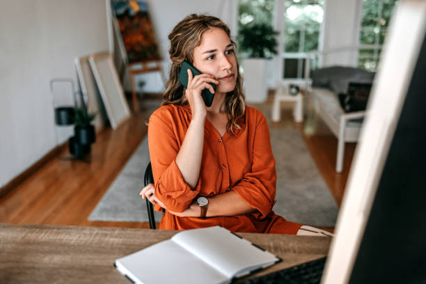 Young woman talking on smart phone at home office Young woman talking on smart phone while working at home office using phone stock pictures, royalty-free photos & images