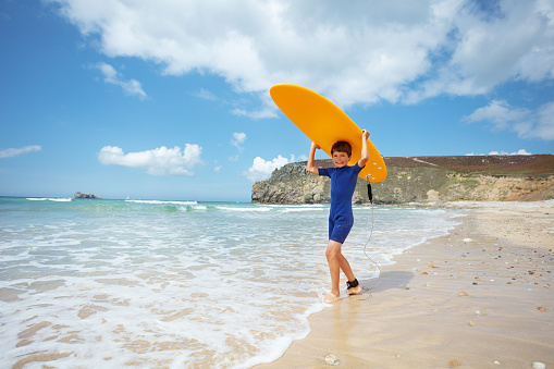 Cute little boy stand holding orange surfboard over head in waves on sand sea beach and smile
