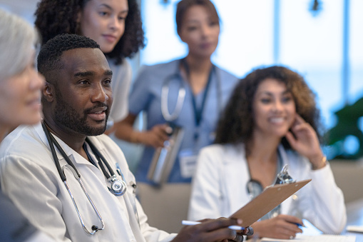 A large group of Doctors, nurses, and other professional medical staff sit closely together as they review a patients case and try to collectively come up with a plan of care.  They are each dressed professionally and focused on the task.