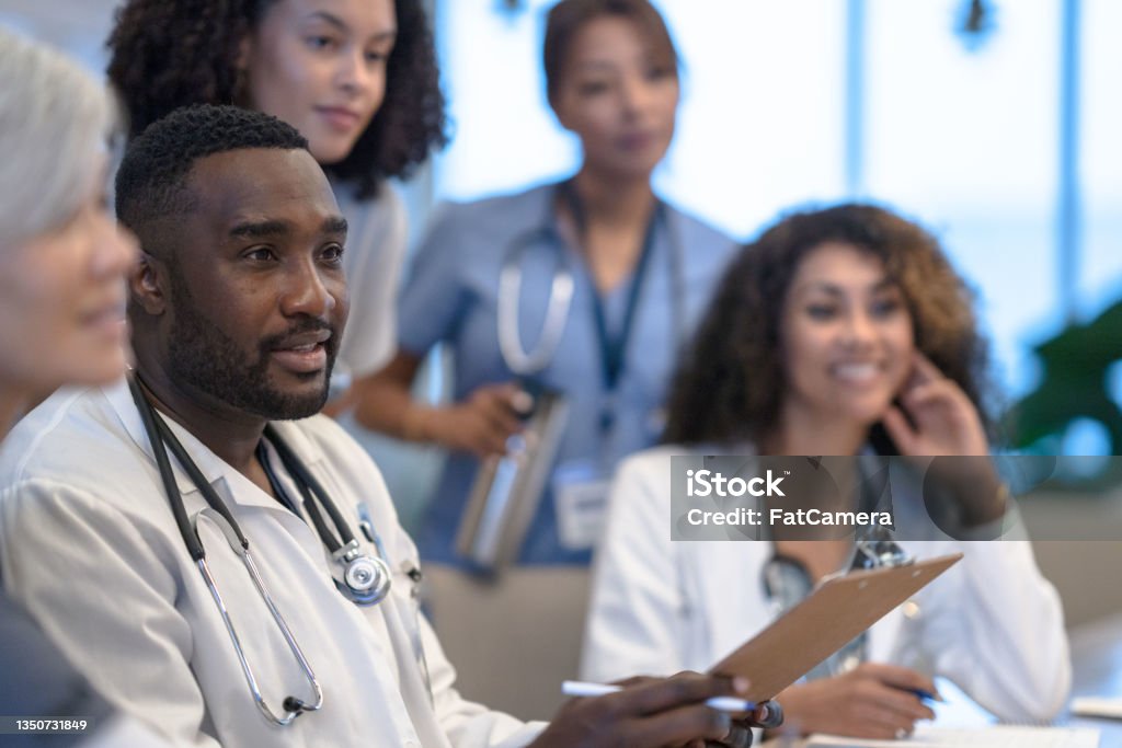 Diverse Medical Team Meeting A large group of Doctors, nurses, and other professional medical staff sit closely together as they review a patients case and try to collectively come up with a plan of care.  They are each dressed professionally and focused on the task. Doctor Stock Photo