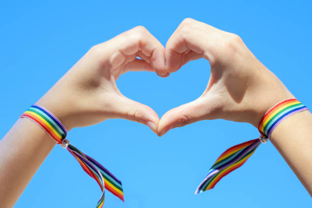 primo piano delle mani della donna in un braccialetto arcobaleno che crea una forma a forma di cuore su sfondo blu del cielo. foto di alta qualità. - gay pride wristband rainbow lgbt foto e immagini stock