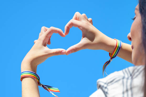 primo piano delle mani della donna in un braccialetto arcobaleno che crea una forma a forma di cuore su sfondo blu del cielo. foto di alta qualità. - gay pride wristband rainbow lgbt foto e immagini stock