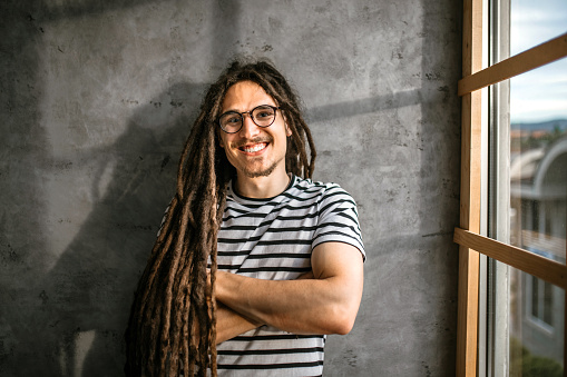 Portrait of young handsome smiling casual businessman with dreadlocks leaning gray concrete wall with arms crossed