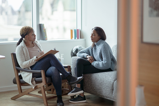 A Therapist meets with her female client in her office.  The client is seated on a sofa with her arms across her body as she looks visibly nervous.  The Therapist is seated in a chair in front of her as she talks about what to expect from the appointment and takes notes on her clipboard.