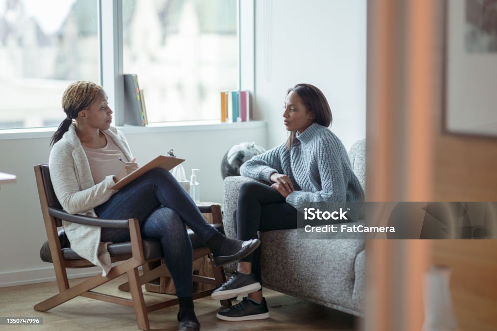 Therapist Meeting with a Client A Therapist meets with her female client in her office.  The client is seated on a sofa with her arms across her body as she looks visibly nervous.  The Therapist is seated in a chair in front of her as she talks about what to expect from the appointment and takes notes on her clipboard. Mental Health Stock Photo
