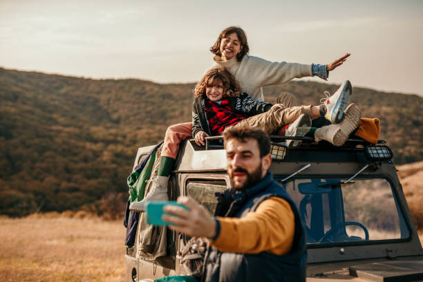 Let’s stop and remember this moment Spontaneous and beautiful photo of a cute family enjoying their adventure, camping and hiking outdoors. They’ve stopped on a beautiful landscape where a beautiful brunette, a mother, is sitting with their sun on the top of the truck, and smiling for the camera, while a father is taking a selfie of all of them. Everyone smiling, stylishly, dressed in colors family camping stock pictures, royalty-free photos & images