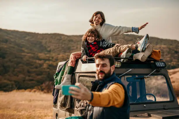 Spontaneous and beautiful photo of a cute family enjoying their adventure, camping and hiking outdoors. They’ve stopped on a beautiful landscape where a beautiful brunette, a mother, is sitting with their sun on the top of the truck, and smiling for the camera, while a father is taking a selfie of all of them. Everyone smiling, stylishly, dressed in colors