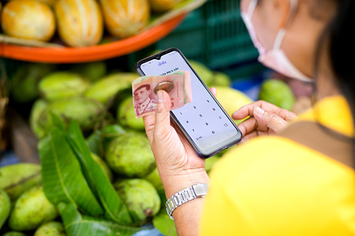 Hand of mature thai woman is holding paper notes and mobile in same hand  and is standing at market stalls for fruits and vegetables in street on local market in Bangkok. Woman is wearing face mask. Rear view capture with screen needed to be unlocked for using app