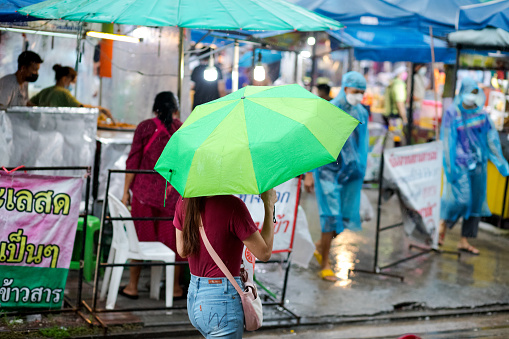 Side view of young thai woman walking with green umbrella in rain and street in Bangkok Chatuchak in late afternoon. In background is a local market