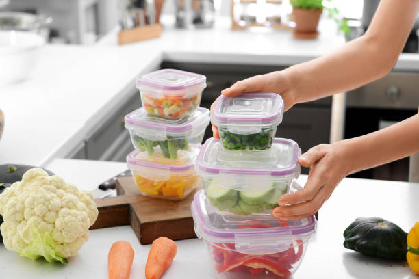 Woman holding stack of plastic containers with fresh vegetables for freezing at table in kitchen stock photo