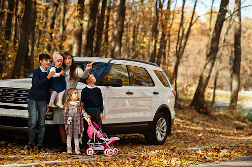 Mother stand near white suv car with four kids in autumn park. Family walk in fall forest.