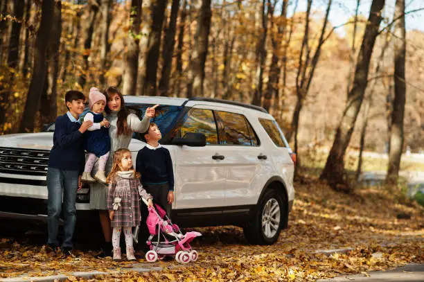 Photo of Mother stand near white suv car with four kids in autumn park. Family walk in fall forest.
