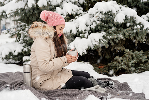Merry Christmas and happy New Year. Beautiful woman in warm winter clothes holding mug drinking hot tea or coffee outdoors in snowy day