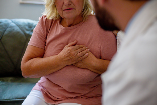 A senior female patient is holding her chest, complaining about pain in her chest.  The doctor is sitting opposite her, and listening about her issues.  Selective focus.