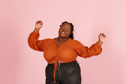 Excited young black plus size body positive woman with dreadlocks in orange top dances standing on light pink background in studio closeup