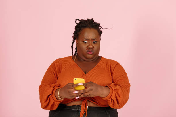 Wondered young black woman holds smartphone on pink background stock photo