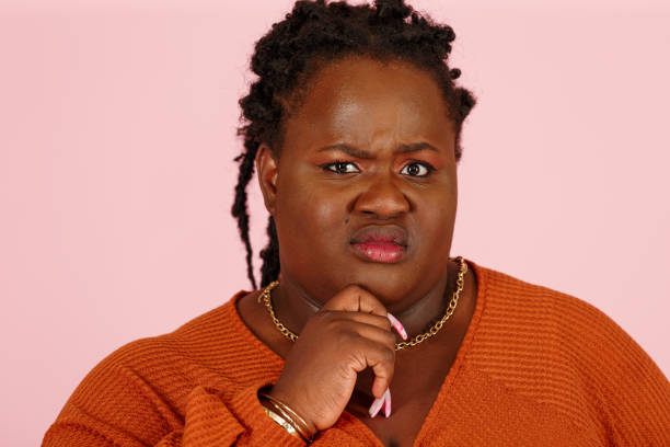 Skeptical black plus size woman touches chin on pink background stock photo