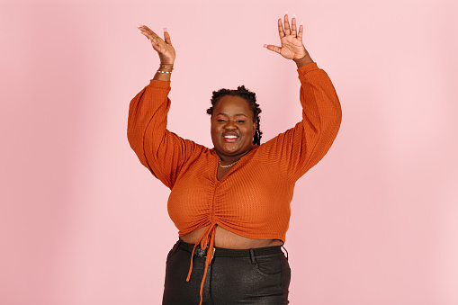 Excited young black plus size body positive woman with dreadlocks in orange top dances standing on light pink background in studio closeup