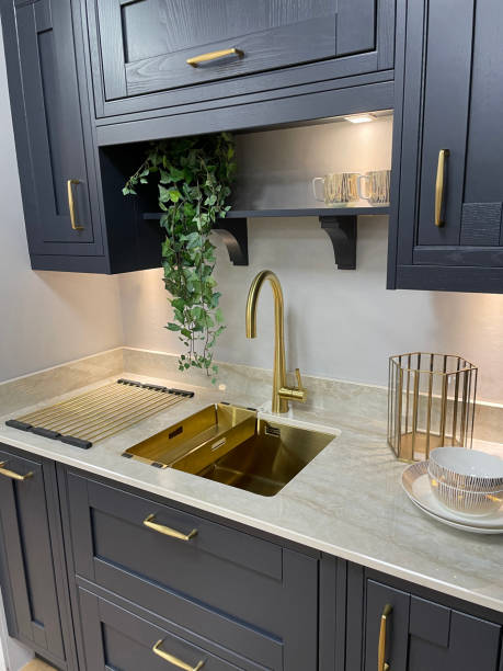 Image of modern kitchen with  navy, wood grain effect wall and floor cabinets, drawers with gold tone handles, white marble kitchen counter inset with rectangular, gold sink with gold, single lever monobloc tap, under cupboard lighting Stock photo showing a gold sink with gold, single lever monobloc tap inset in white marble kitchen counter over navy, wood grain effect floor cabinets. red kitchen cabinets stock pictures, royalty-free photos & images
