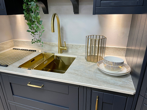 Stock photo showing a gold sink with gold, single lever monobloc tap inset in white marble kitchen counter over navy, wood grain effect floor cabinets.