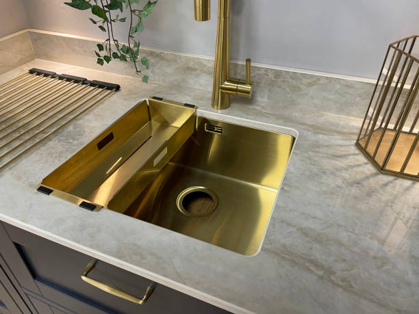 istock Close-up image of modern kitchen with rectangular, gold sink with gold, single lever monobloc tap inset in white marble kitchen counter, navy, wood grain effect floor cabinets, drawers with gold tone handles 1350795371