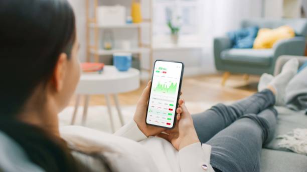 Young Woman at Home Sitting on a Couch and Using Smartphone with Stock Market App to Check Share Prices. Graph is Going Up Making a Great Profit. In the Background Cozy Homely Atmosphere. mujer joven en casa sentada en un sofá y usando un teléfono inteligente con la aplicación del mercado de valores para verificar los precios de las acciones. el gráfico está subiendo obteniendo una gran ganancia. en el fondo acogedor ambiente hogareño