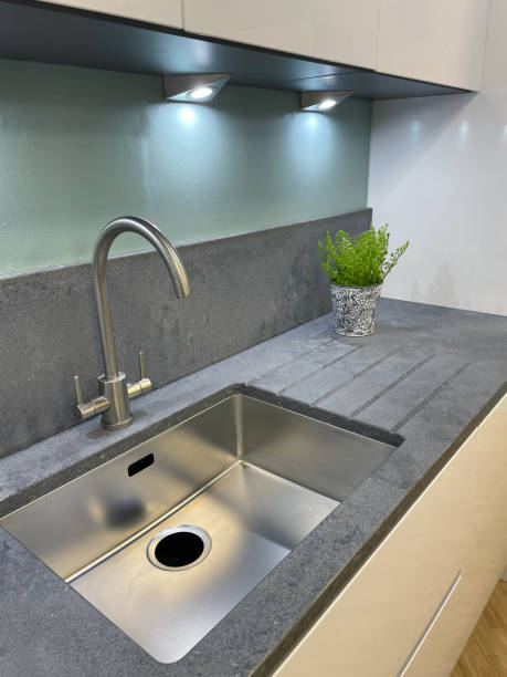 istock Image of stainless steel sink with grey granite worktop, integrated granite draining board with grooves, runnels and drains, concealed under wall cabinet lights, potted plant 1350796066