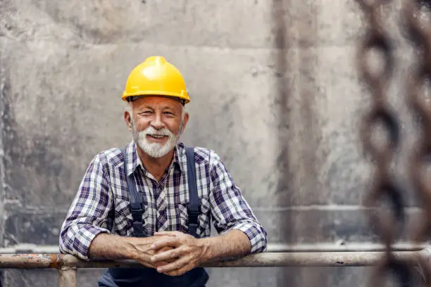 A smiling, senior worker with a helmet on his head, leans on the railing in the factory and takes a break from work in heavy industry. A senior worker in a factory.