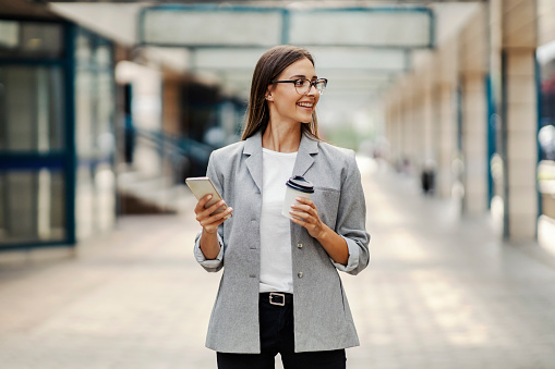 A happy, elegant, young businesswoman standing in the business center, holding takeaway coffee and using the telephone. Businesswoman on a coffee break