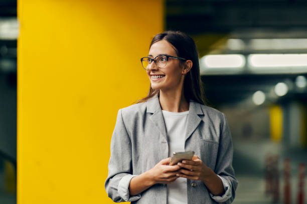 una joven feliz y elegante está parada en el estacionamiento y usa su teléfono para escribir un correo importante. una mujer de negocios que usa tecnologías en el exterior - hora punta temas fotografías e imágenes de stock