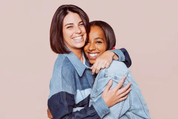 Image of two cheerful multinational girls hugging and smiling together at camera. Caucasian and mixed race woman portrait. Copy space.