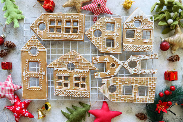 istock Image batch of homemade, house-shaped, gingerbread biscuits, cookies iced with white glace icing on white, metal cooling rack, Christmas village display surrounded by felt Christmas tree and star-shaped ornaments, marble effect background, elevated view 1350812888