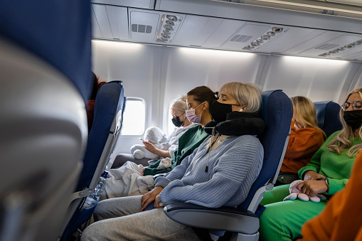 A side-view shot of female friends sitting next to each other on an airplane wearing protective face masks, they are excited to be traveling on holiday.