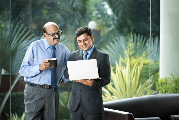 Two businessmen laughing while drinking coffee and using laptop at office