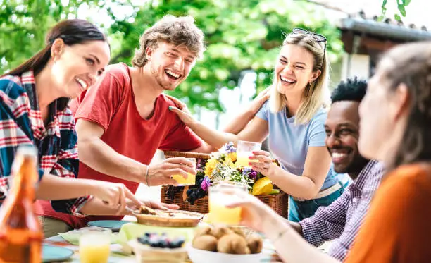 Photo of Happy young men and women toasting healthy orange fruit juice at farm house picnic - Life style concept with alternative friends having fun together on afternoon relax time - Bright vivid filter