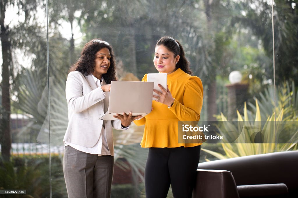 Two businesswomen using laptop and digital tablet at office Two happy businesswomen discussing using laptop and digital tablet at office India Stock Photo