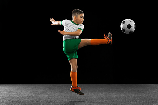 Making a toe punt. Portrait of boy, child, football player in uniform training, kicking ball isolated over black background. Concept of action, team sport game, energy, vitality. Copy space for ad.
