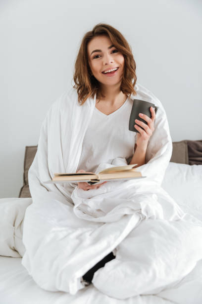 istock Lady sitting on bed while reading book and drinking coffee. 1350830711