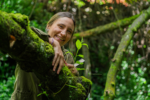 Young Latina woman from Bogota Colombia between 20 and 29 years old looks at the camera in a portrait as she leans against the tree she found in the forest while reading