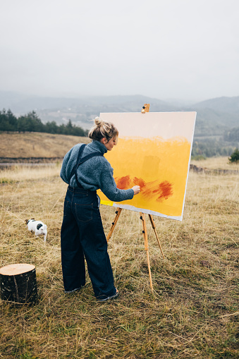 Serious woman artist with palette and brush painting an art canvas outdoors with her dog