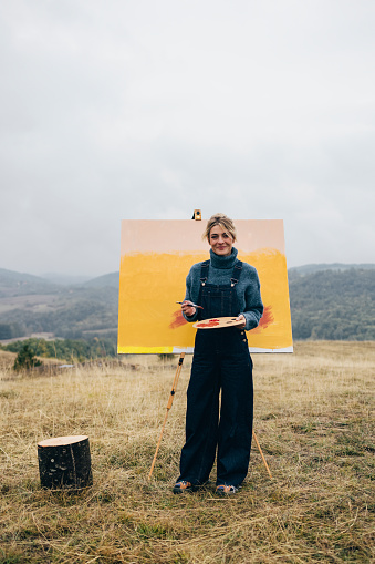 Happy woman artist with palette and brush painting an art canvas outdoors and looking at camera