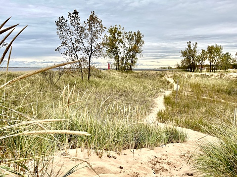 Sand dune and footpath in the foreground look to the horizon where the moody sky meets Lake Michigan. Red lighthouse in the distance. Surrounded by wild grass and trees.
