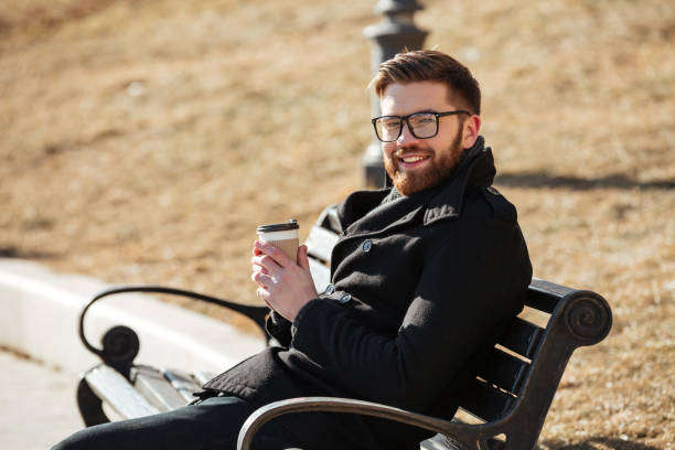 istock Cheerful bearded young man sitting on bench and drinking coffee 1350834518