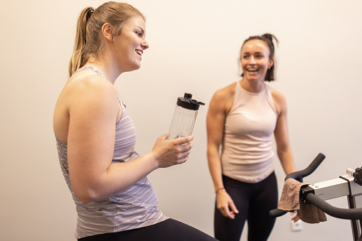Woman exercising on a stationary bike smiling with her personal trainer. Female sitting on workout bicycle drinking water from a bottle and talking with her fitness trainer in gym. Woman with trainer taking break after fitness training at gym.